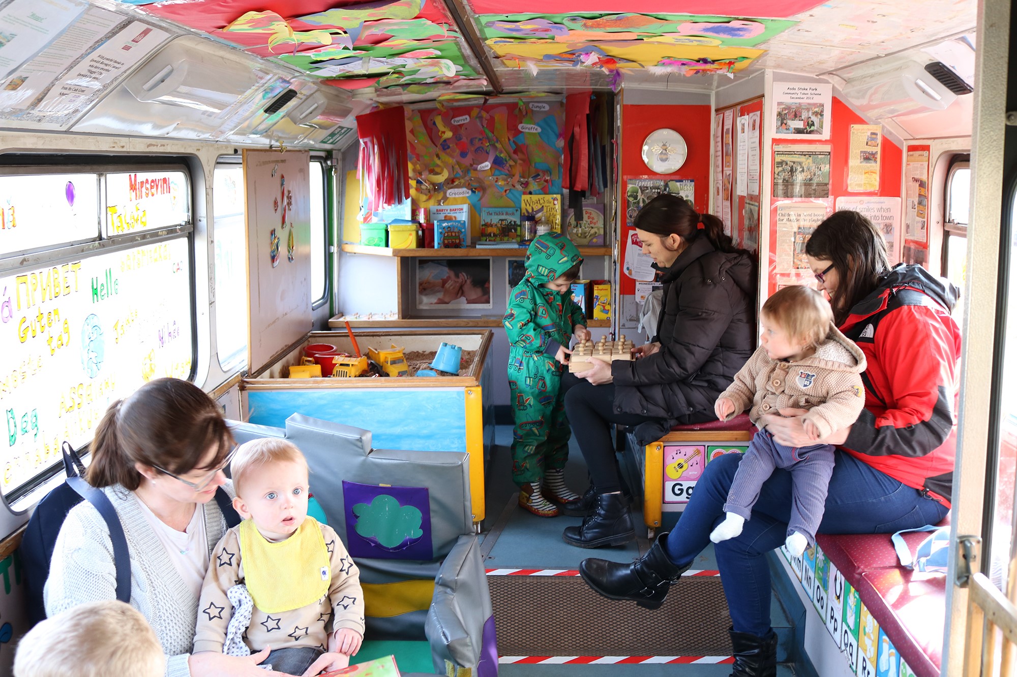 Three women with young children sit inside the bus engaging in various activities. There is a sand pit, posters and children's artwork on the walls and ceiling.