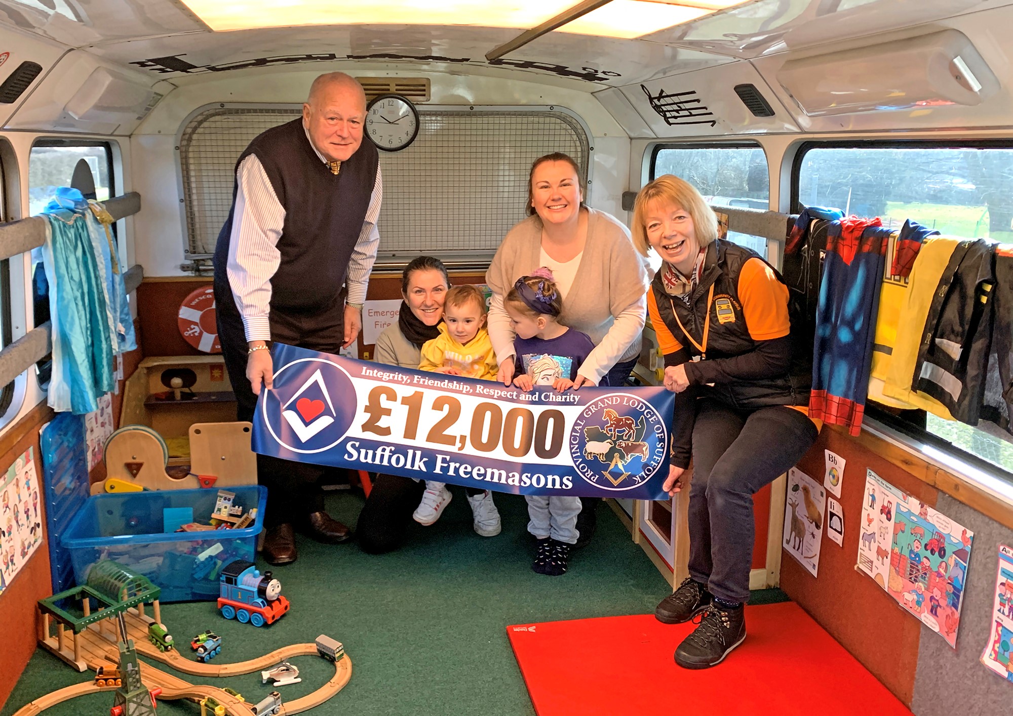 A group of people; An older man, three women and two young children, sit inside the bus holding the '£12,000' banner and smiling.