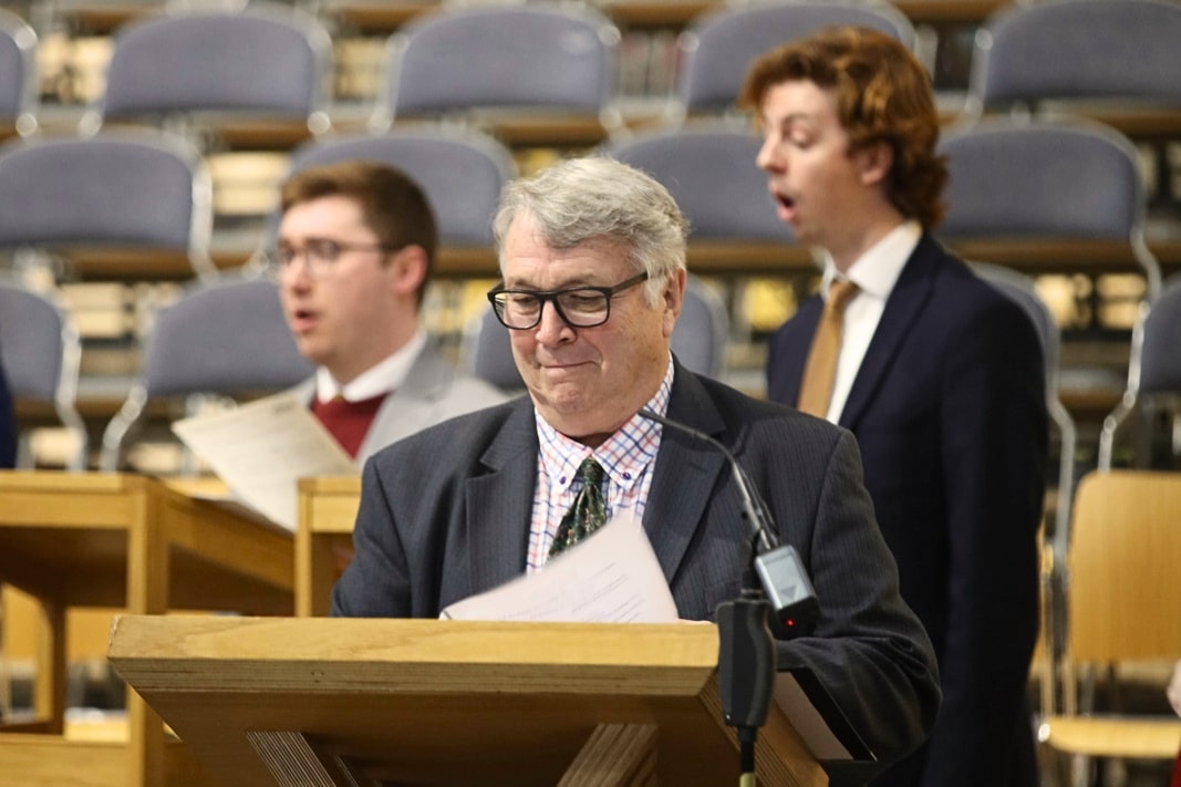 An image of three carol singers. An older man in focus and two younger men behind out of focus.