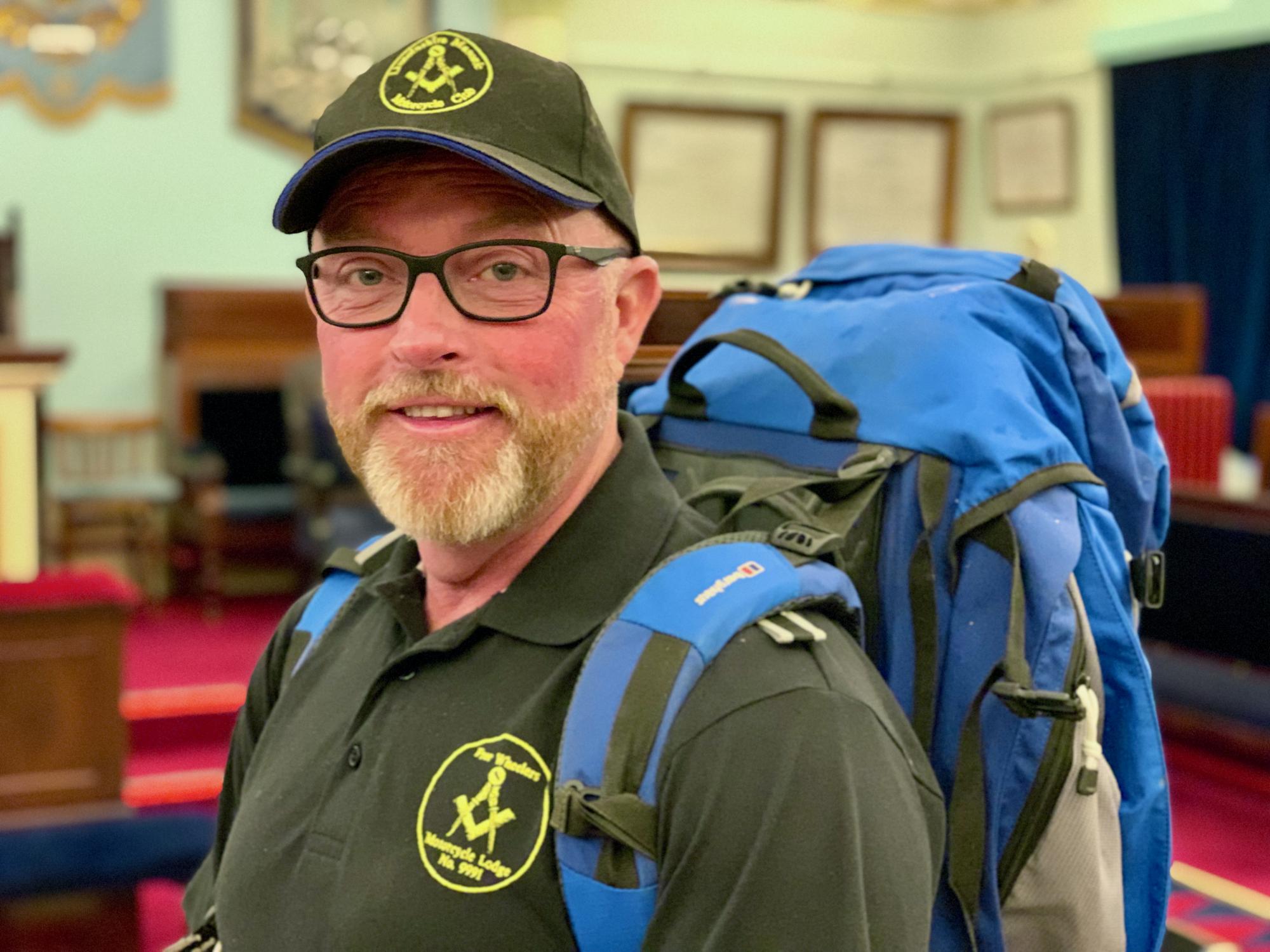 A bearded man with classes wearing a green cap and polo shirt smiling at the camera. He is wearing a blue backpack.