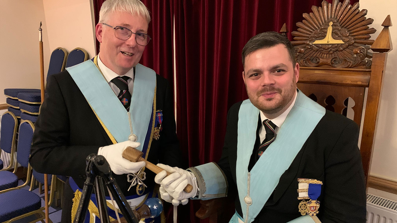 A father and son in freemasonry regalia. The younger man has short dark hair and stubble, he is sat in an ornate chair. The older man has grey hair and glasses and is stood to the right of the son. They are both holding onto a wooden gavel.