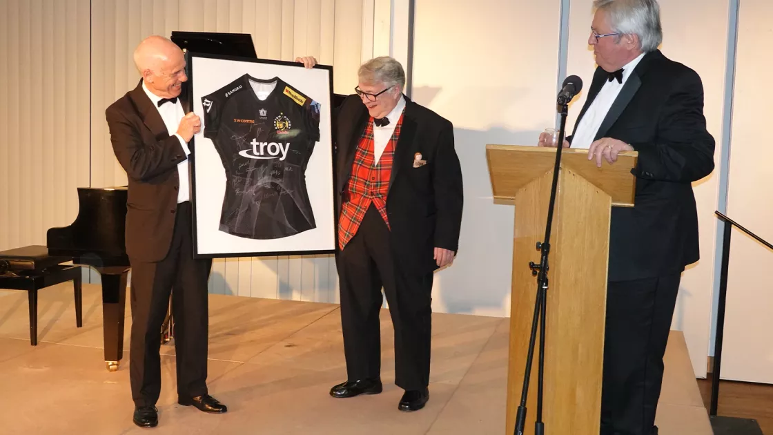 two men in formal wear holding a framed Rugby shirt. A third man stands at a podium.